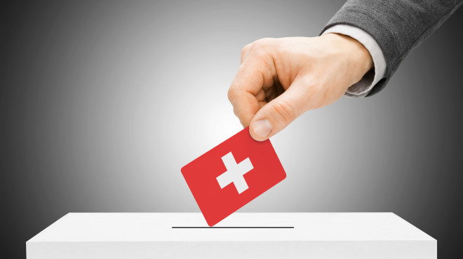 Männliche Hand legt roten Wahlzettel mit Schweizerkreuz in Wahlurne