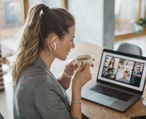 Frau sitzt mit einem Kaffee vor einem Laptop auf dem ein Online-Meeting stattfindet